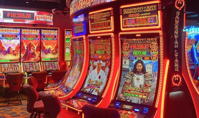 Close-up of brightly lit slot machines in a casino, including Phoenix Link and Buffalo Ultimate Stampede games with jackpot amounts displayed.